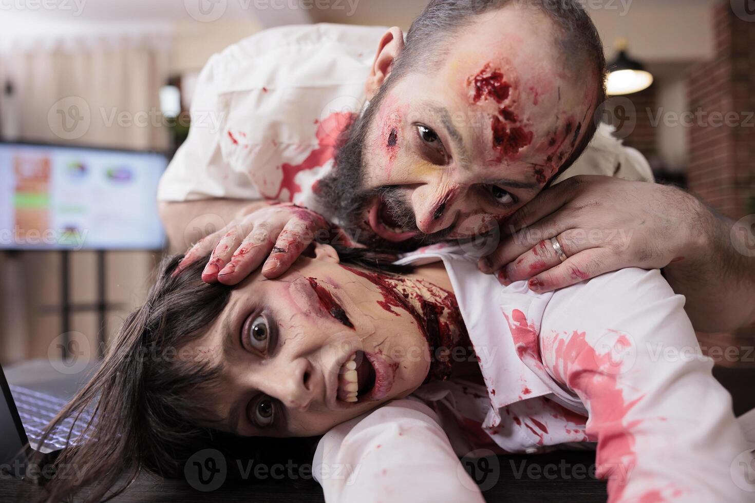 Colleagues having fun in office dressed as zombies for Halloween event, taking selfies. Man and woman covered in fake bruises pretending to be undead creatures using selfie camera to take photos