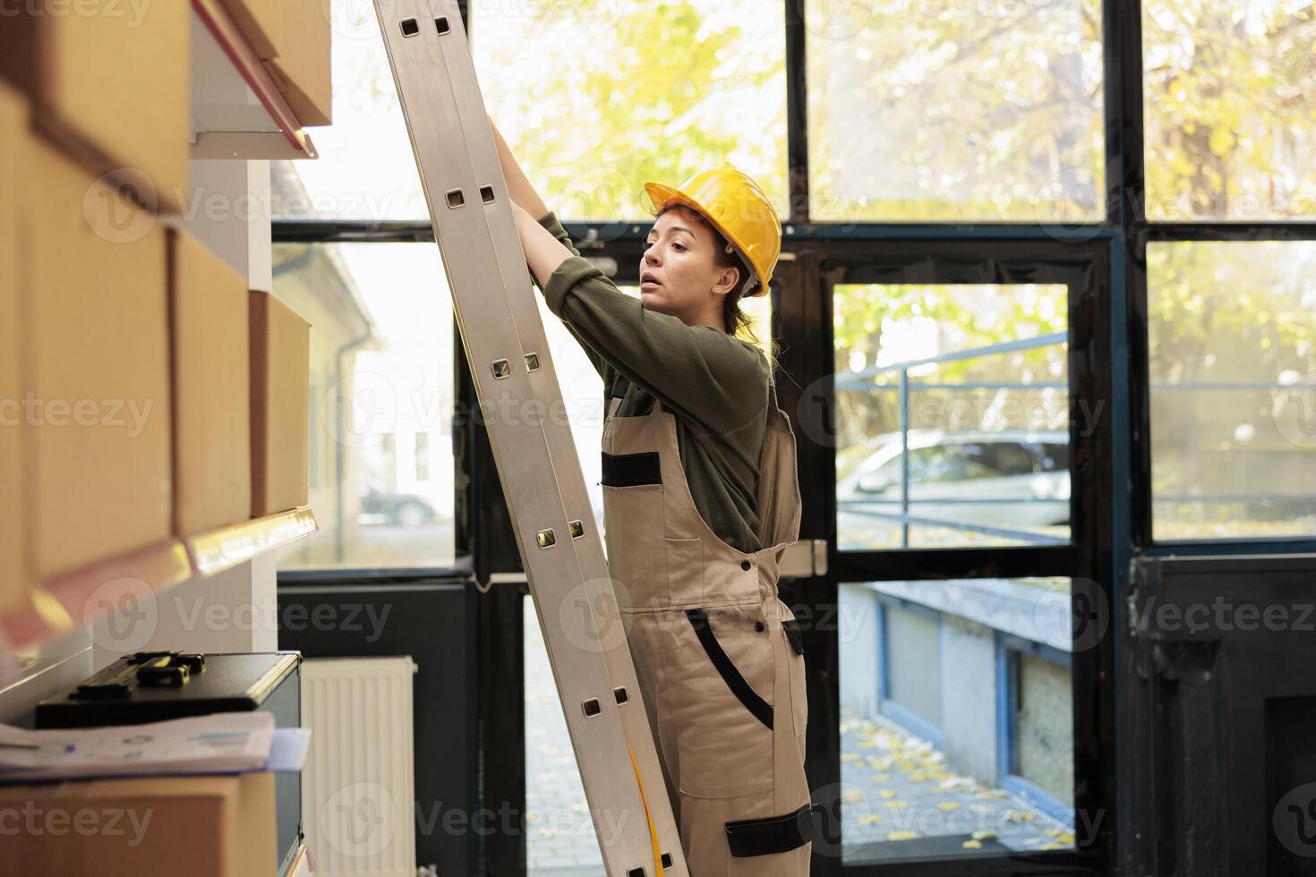 Storage room manager standing on ladder in warehouse, checking cardboard boxes full with merchandise. Supervisor woman working at customers orders before preparing delivery in storehouse photo