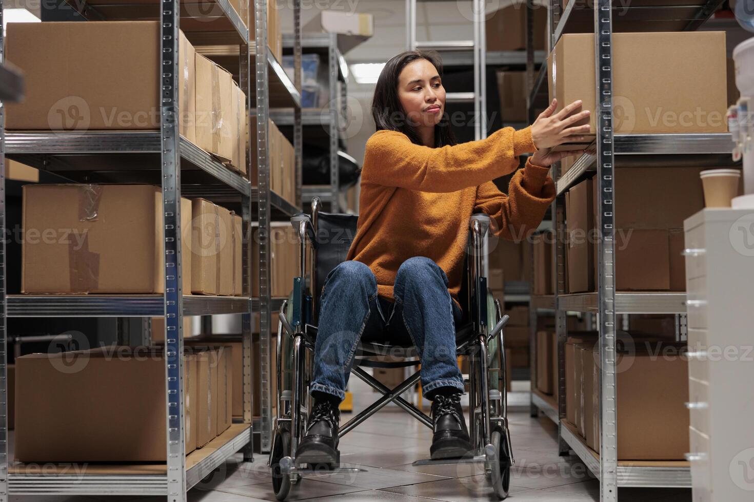 Woman wheelchair user taking cardboard box from shelf to prepare customer order for dispatching in warehouse. Asian package handler holding parcel, working in disability friendly storehouse photo