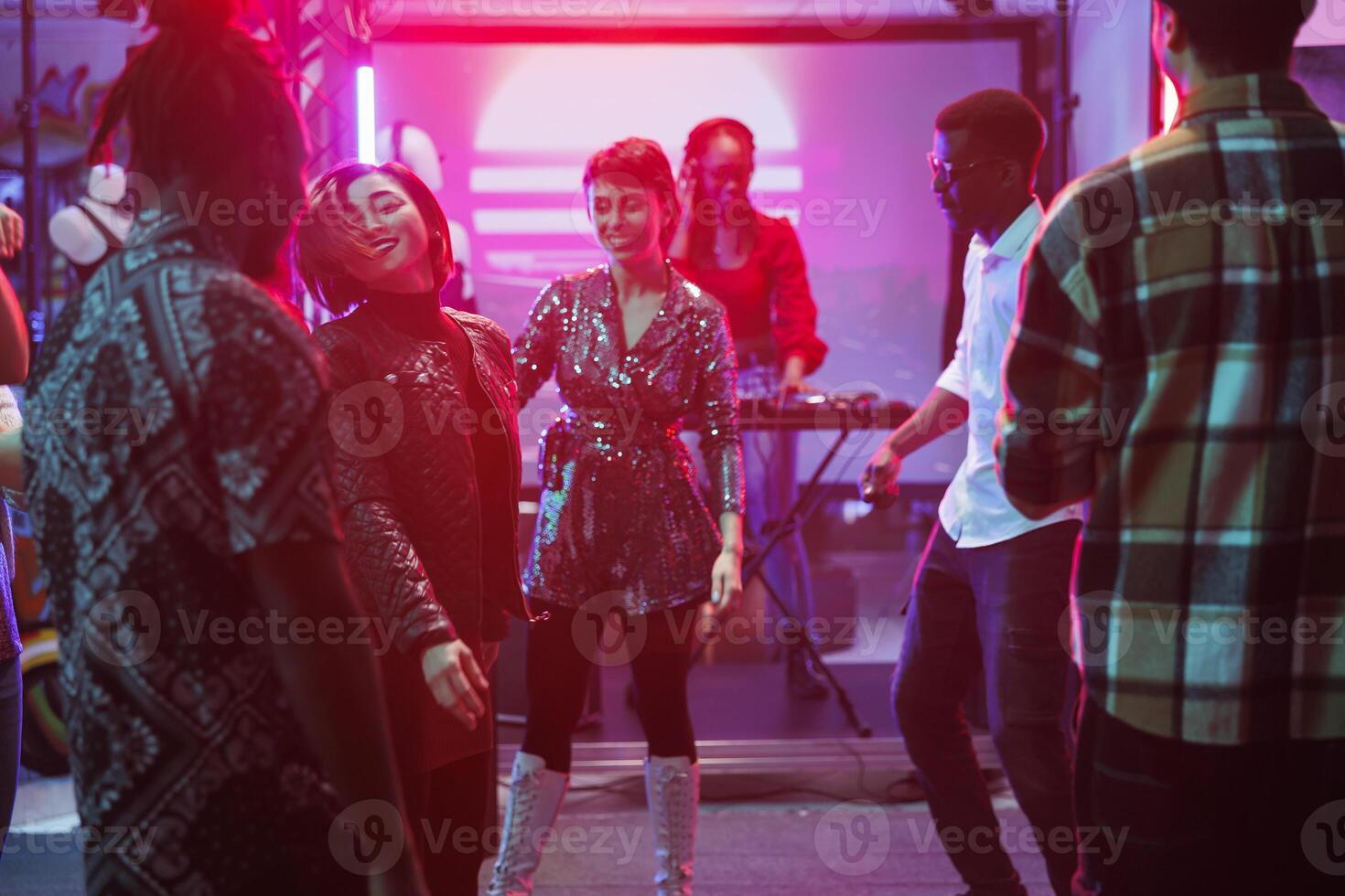 People dancing and enjoying live electronic music show in nightclub. Young diverse clubbers crowd moving on dancefloor while dj performing on stage with spotlights in club photo