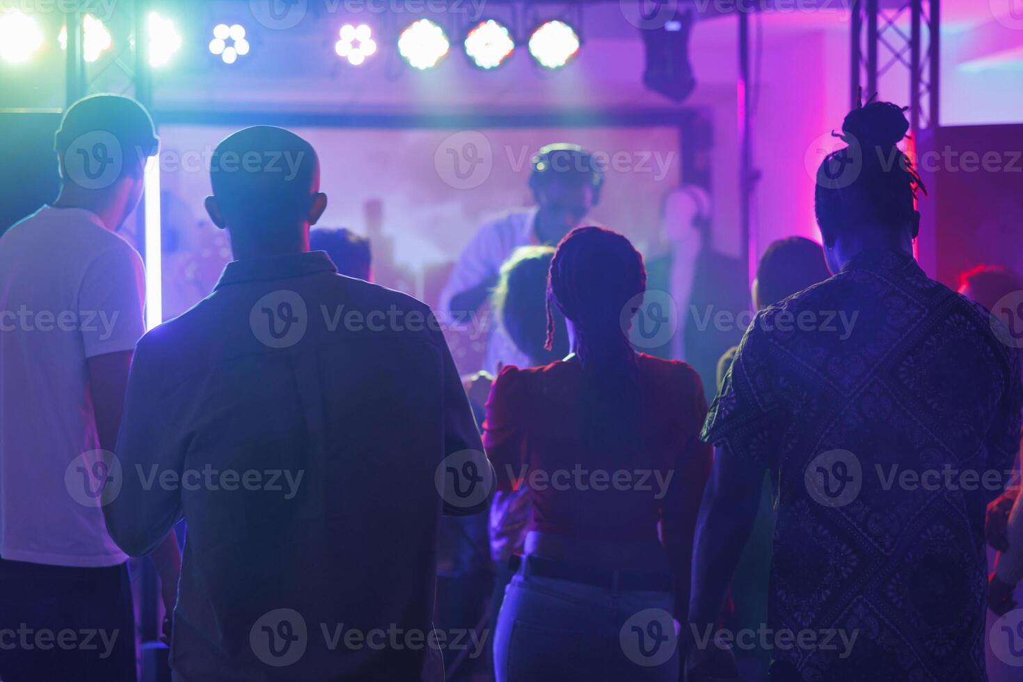 People partying and attending dj performance at nightclub discotheque. Young diverse men and women standing on crowded dark dancefloor while musician playing on stage in club photo