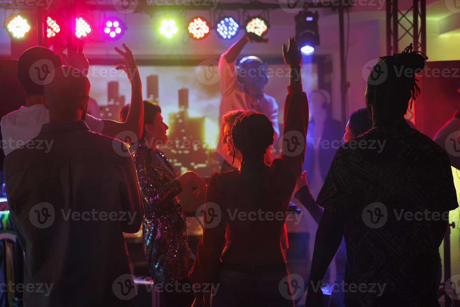 Clubbers partying and dancing while attending dj performance in nightclub. Young people crowd standing with raised hands on dancefloor illuminated with spotlights at discotheque photo