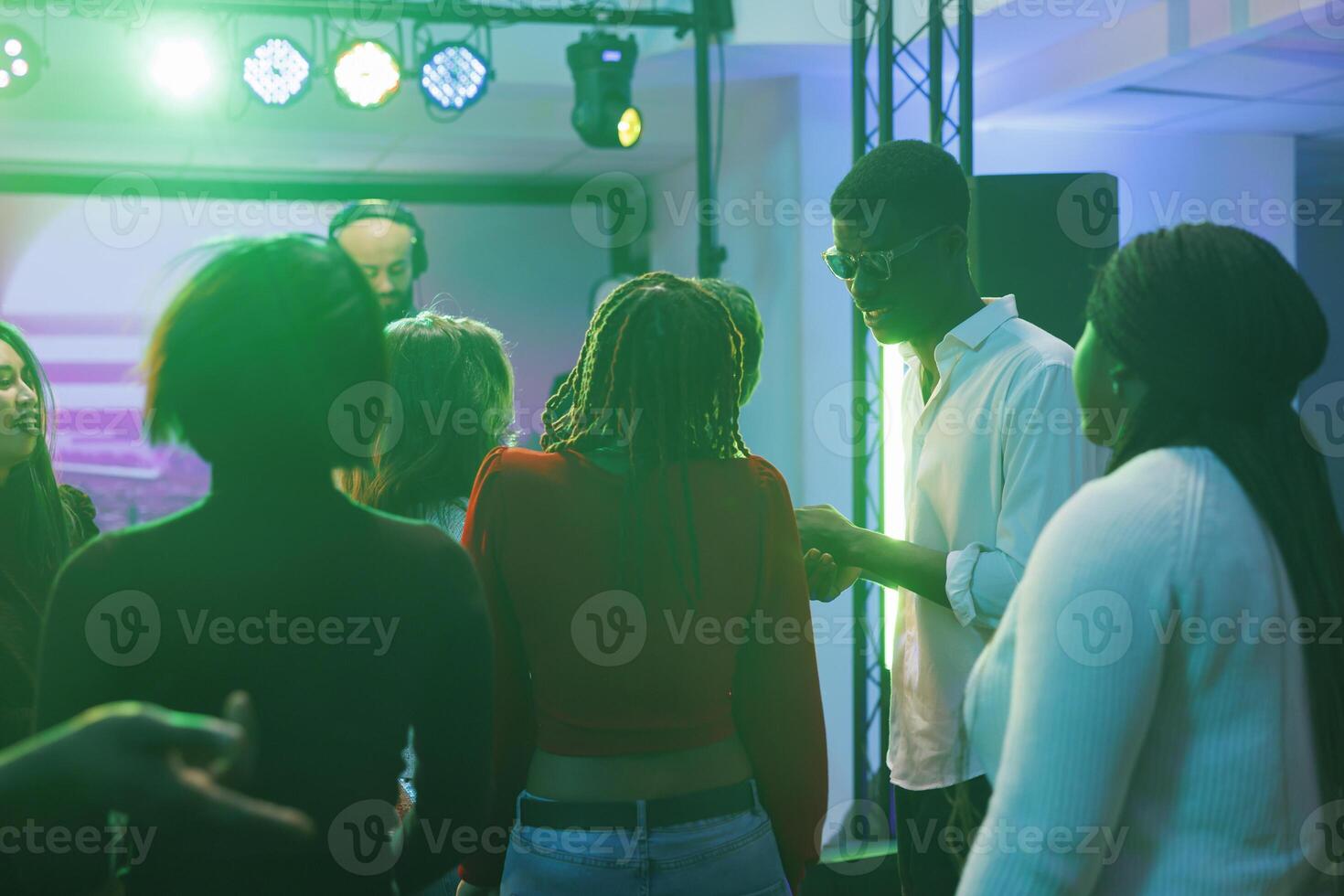 People partying and having fun at social gathering while attending dj concert in nightclub. Diverse crowd relaxing on dancefloor while musician performing on stage in dark club photo