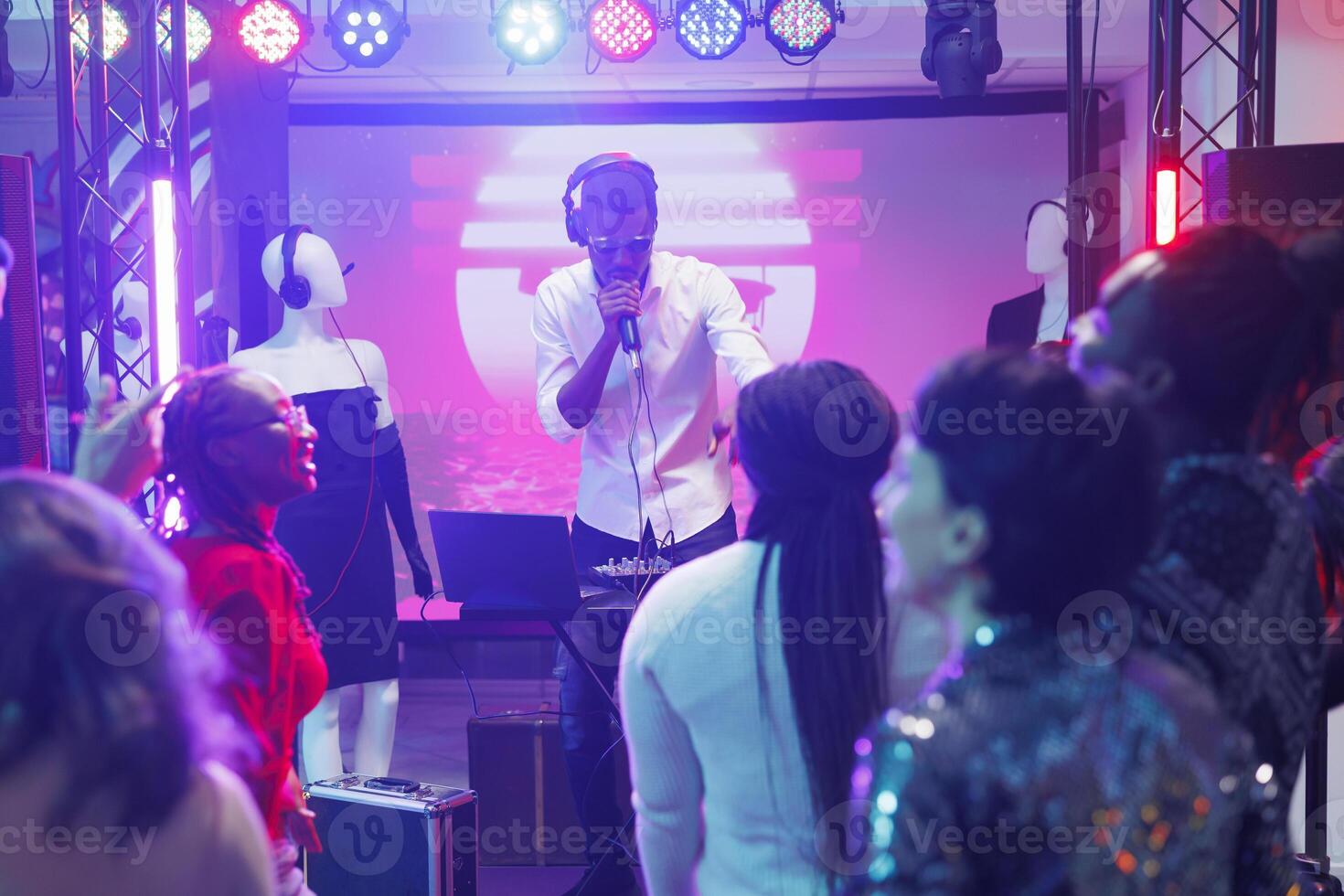 Young man dj mixing music using controller and singing in microphone on club stage with spotlights. African american musician entertaining clubbers crowd at party in nightclub photo