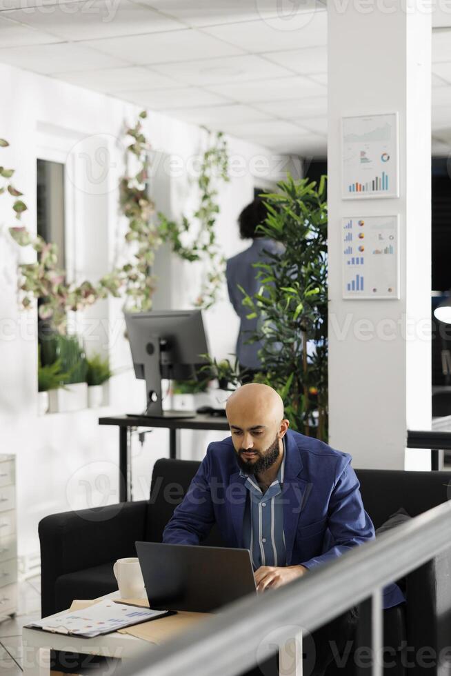 Arab businessman solving marketing strategy problems on laptop while working in start up business office room. Man entrepreneur developing product sales plan on computer in green open space photo