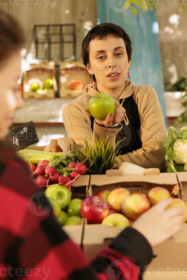 local vendedor vistiendo un delantal y hablando a clientes mientras de venta orgánico Fresco manzanas a un agricultores mercado pararse. un joven mujer sostiene bio natural agricultura productos crecido en la zona. foto