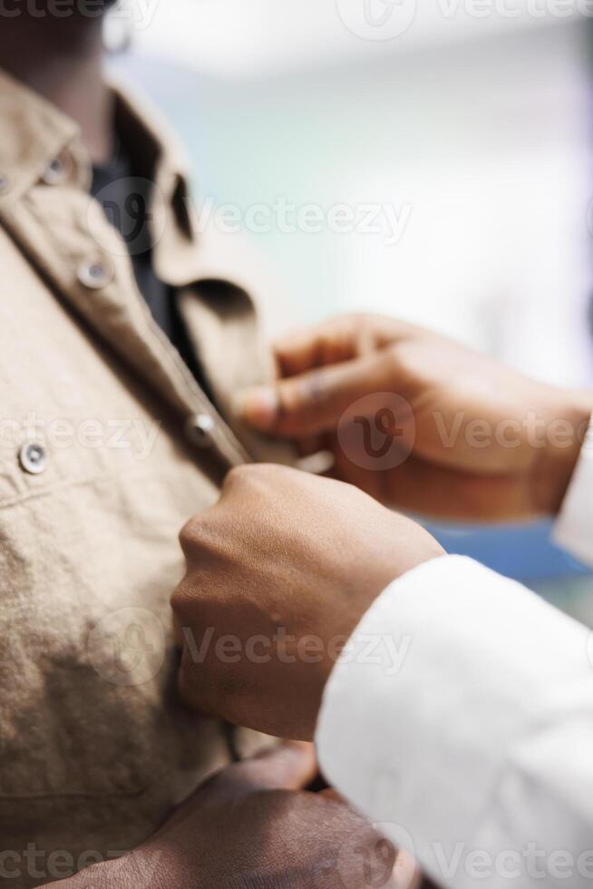 Clothing store assistant hands fastening buttons on customer shirt. African american fashion boutique employee helping client to try on casual apparel before buying with close up on arms photo