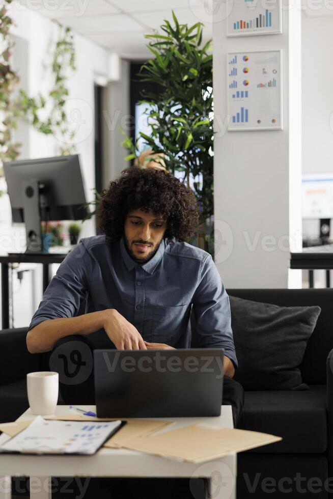 Arab executive manager working at start up project development in business office. Young businessman sitting on couch at workspace and checking marketing research on laptop photo
