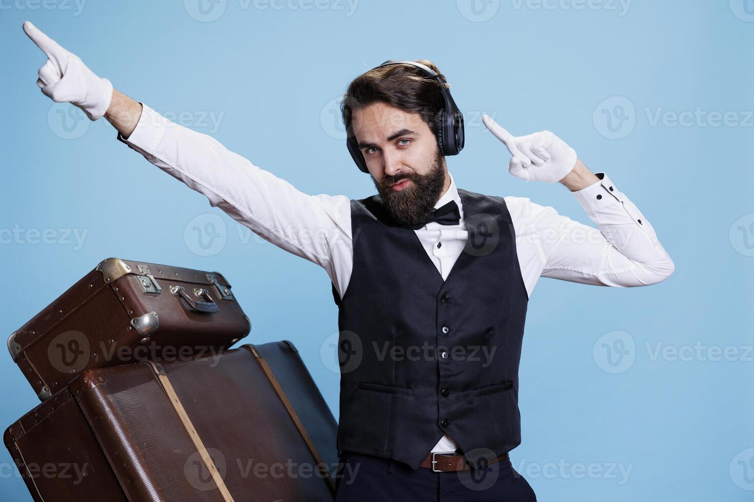 Blue background with enthusiastic bellboy singing and enjoying tunes while he is using audio earpieces. Person in a suit dancing and behaving openly at the camera, having fun with music. photo