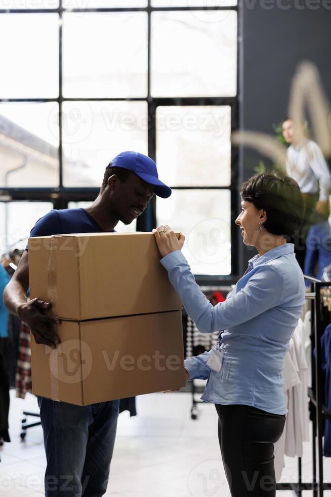 Tienda gerente que se discute cliente pedidos detalles con africano americano mensajero en moderno boutique. hombre vistiendo entrega uniforme, participación cartulina cajas con elegante ropa en compras centrar foto