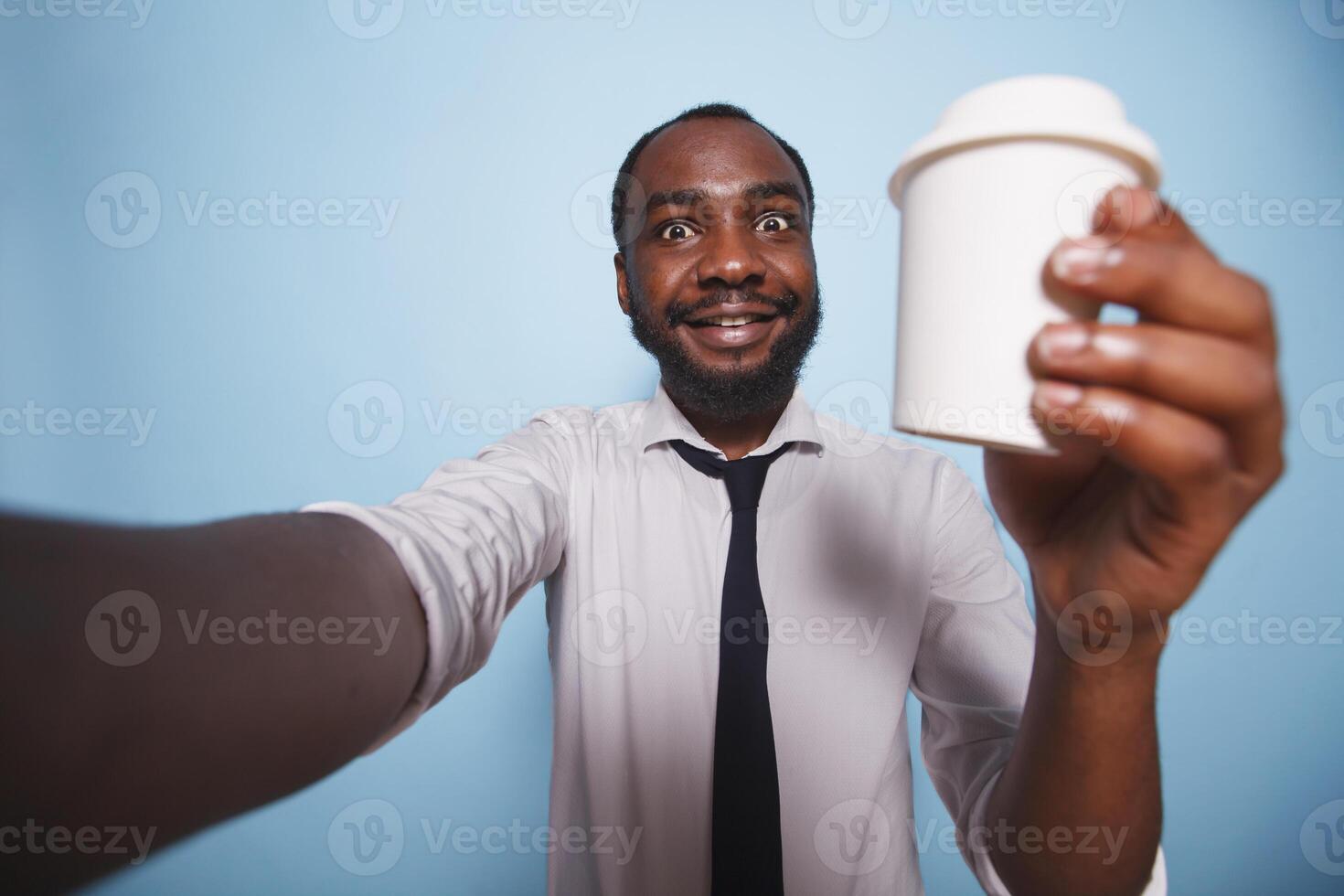 Black man having a call while holding up cup of coffee against isolated background. Smiling african american dressed in white shirt making a selfie photo with beverage container in his hand.