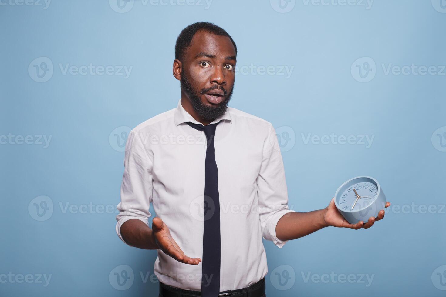 Young businessman looking surprised running out of time holding alarm clock while wearing white shirt and black tie. African american office worker panicking for being late for a meeting. photo