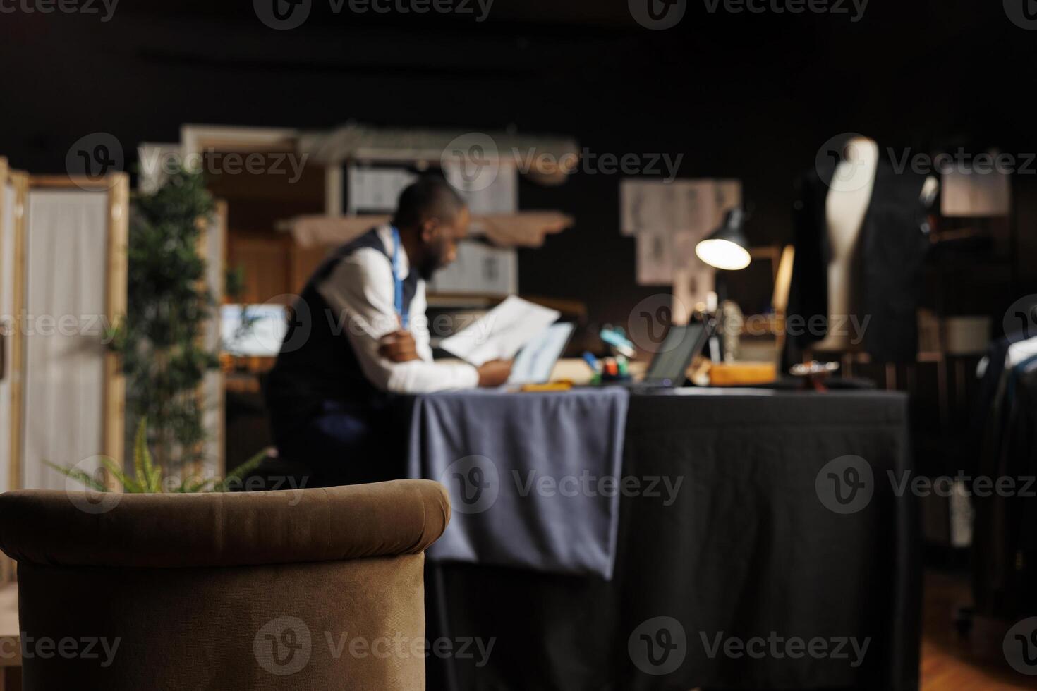 Hip level shot of cozy tailoring studio with blurry african american dressmaker in the background studying sartorial designs. Couturer inspecting sketches of upcoming fashion collection photo
