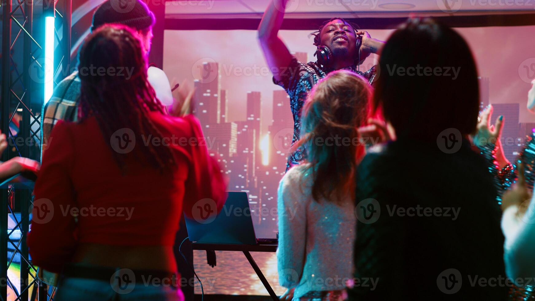 Young man clubbing with people at bar, having fun with DJ mixing station on nightclub stage with disco ball. Funky person creating live performance on audio panel, spotlights at club. photo