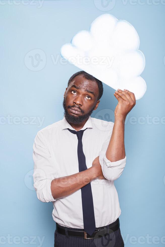 Portrait of male entrepreneur in white shirt looking up at white paper idea cloud brainstorming while standing in front of blue background. African American man thinking under thought bubble. photo