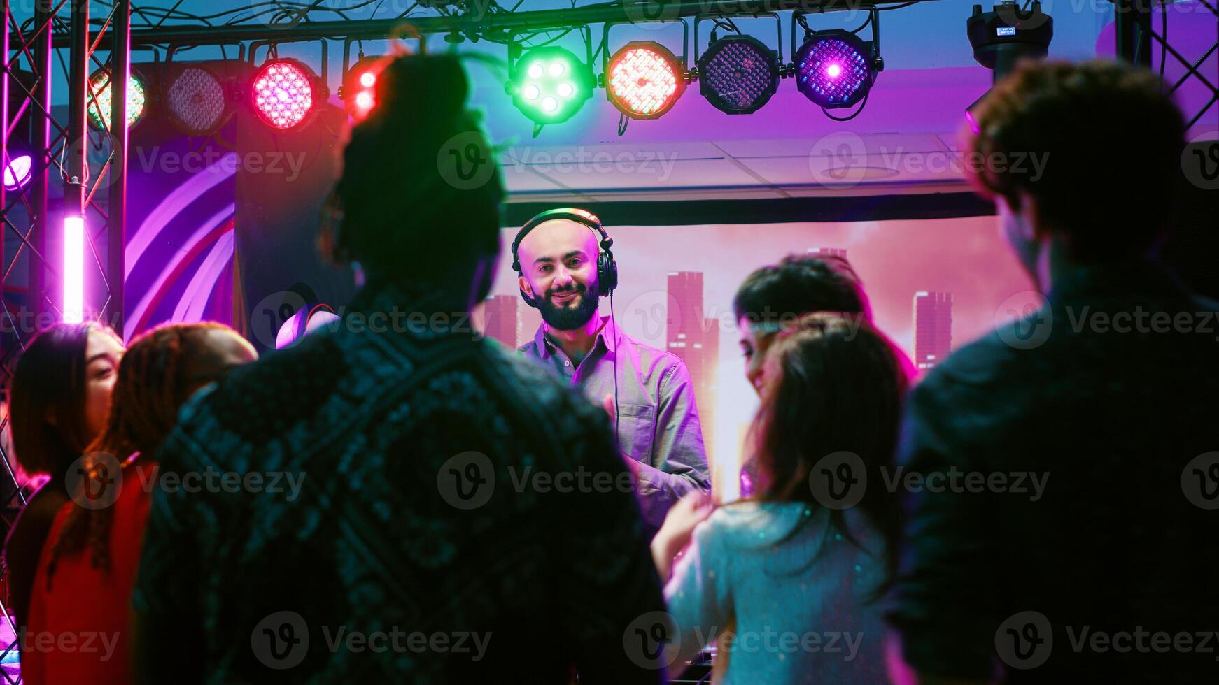 Male DJ putting music at club party, mixing electronic sounds at audio equipment station. Diverse group of friends jumping around and dancing on dance floor, clubbing entertainment. photo