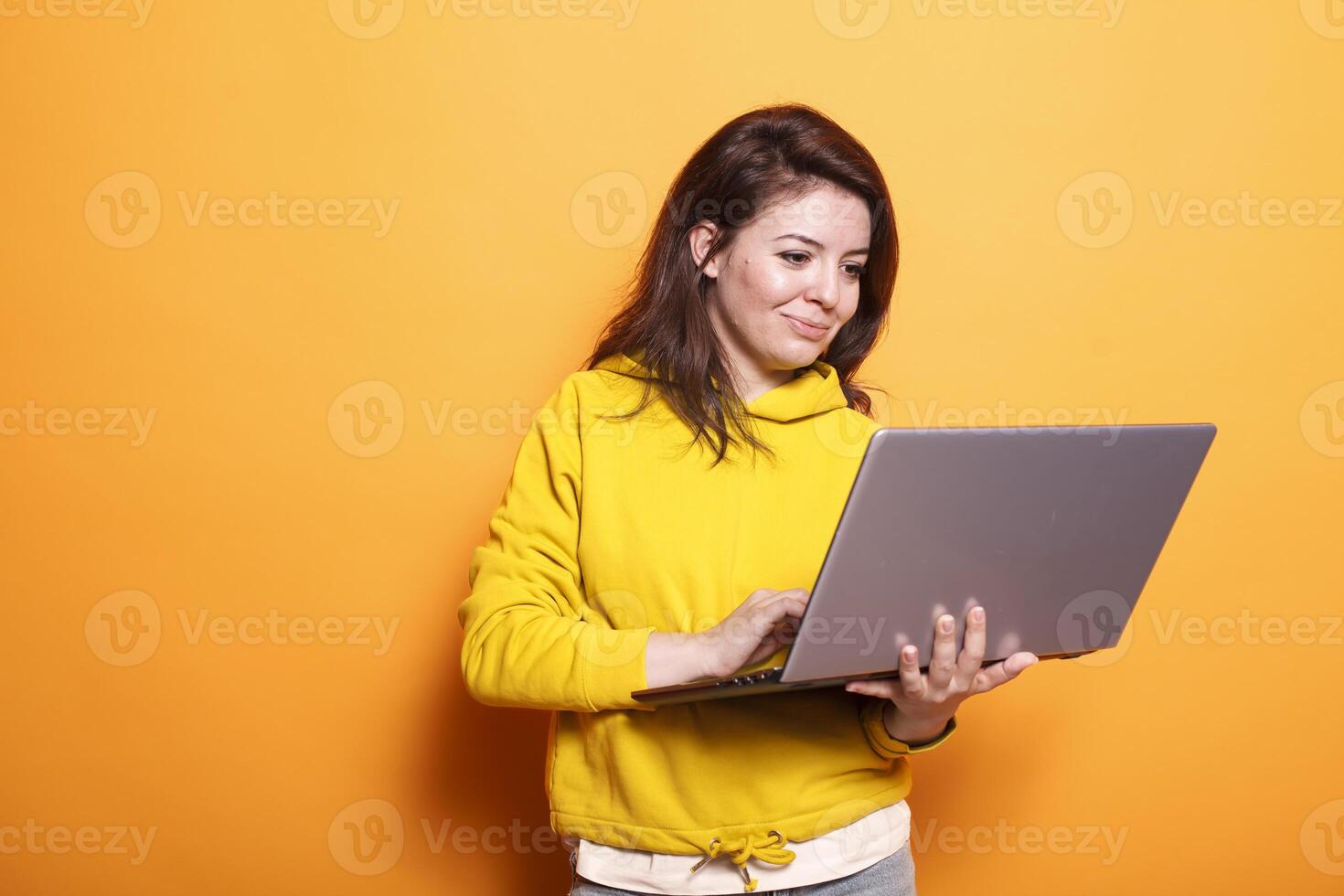 In the studio, young adult looks at a laptop to use technology on camera. Portrait of a modern woman standing over an isolated background, holding a digital device and surfing the internet. photo