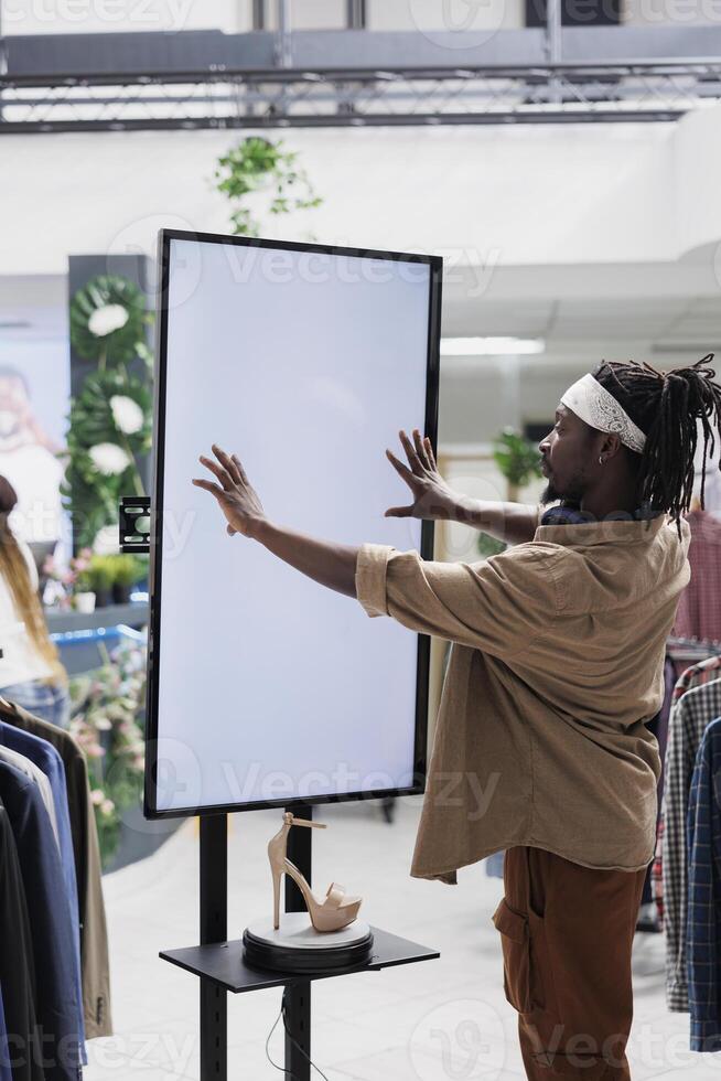 African american man interacting with digital screen offering shoes to customer in shopping center. Clothing store buyer touching white empty display mock up to check stiletto brand options photo