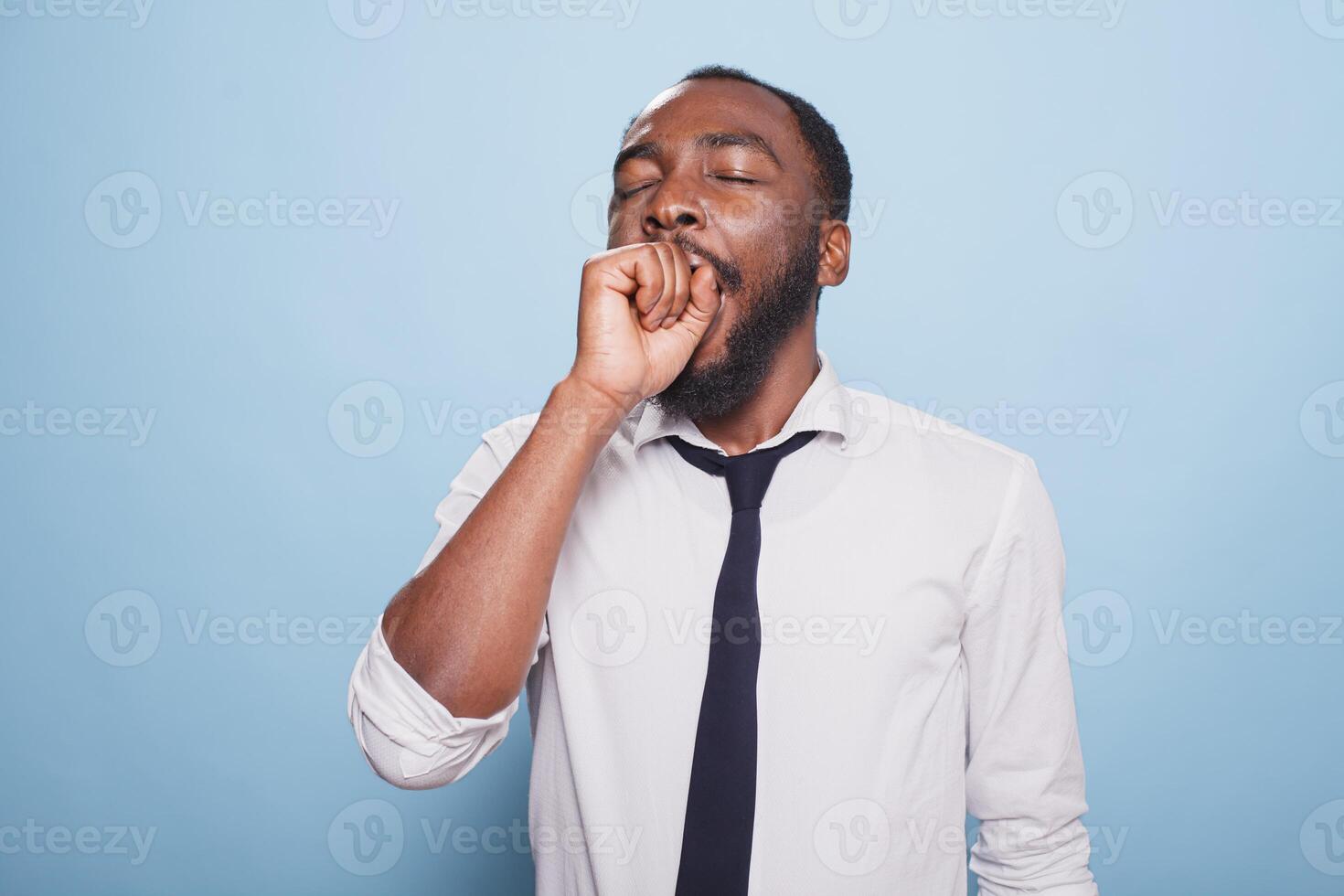 Tired black man yawning with his mouth covered in front of isolated background. Exhausted African American male entrepreneur looking at camera, feeling fatigued and overworked. photo
