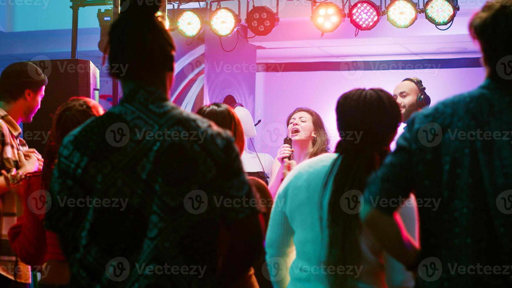 Happy woman doing karaoke at party, singing songs with DJ in front of friends on dance floor. Confident girl having fun with live show perfromance next to electronic mixing station at nightclub. photo