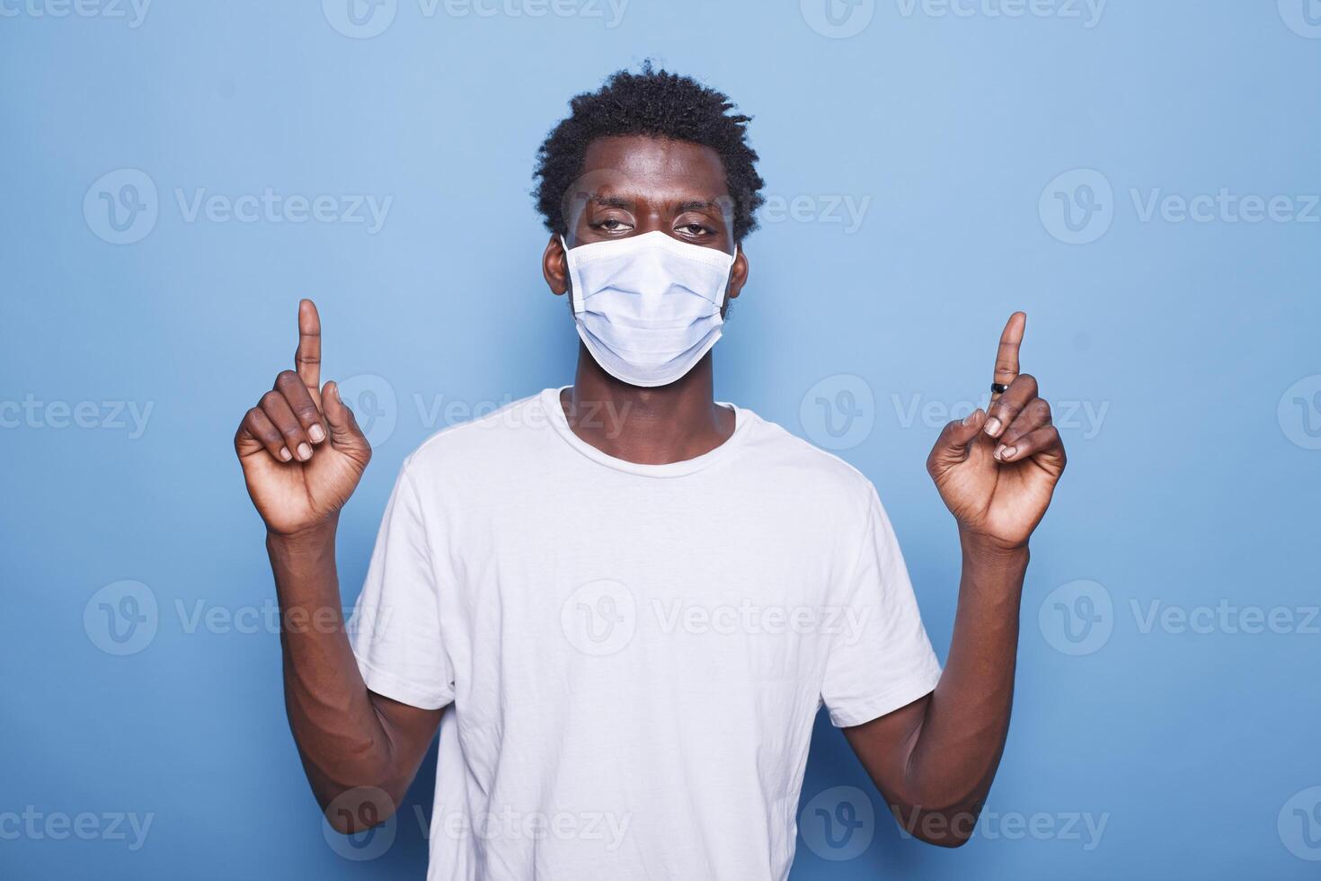 Portrait of black man wearing mask for coronavirus protection is pointing up with his two index fingers. African american guy wearing face mask while doing sign with hands in the studio. photo