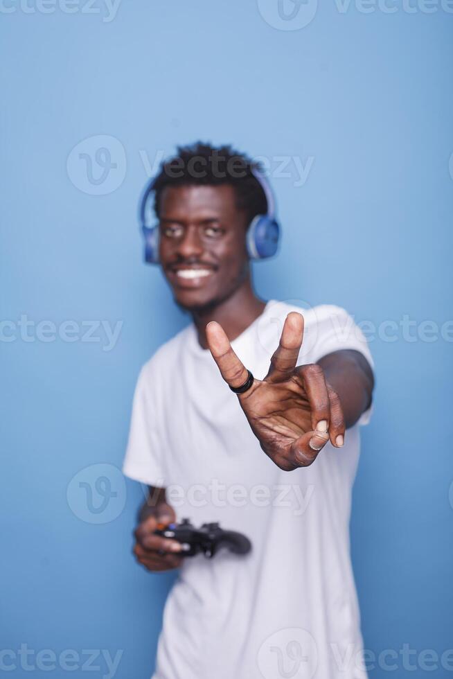 African American man wearing headphones and carrying controller gesture peace sign towards camera. Black person standing in front of isolated backdrop makes happy gesture with their hand. photo