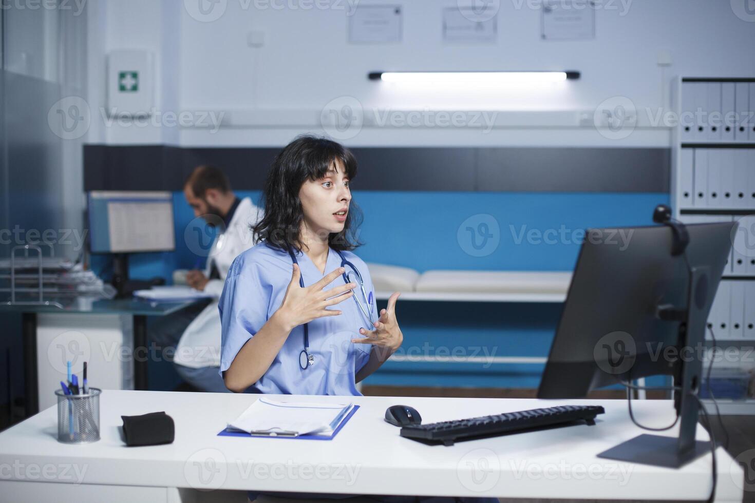 hembra enfermero en un hospital comunica mediante un llamada utilizando su escritorio ordenador personal. caucásico mujer sentado en el clínica oficina teniendo un virtual reunión. colaboración y tecnología en cuidado de la salud. foto