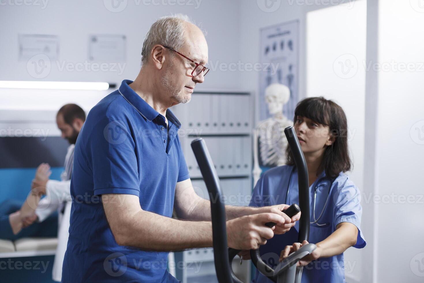 Female medical specialist assisting elderly outpatient on stationary bicycle for physical therapy. Senior man receiving physiotherapy to recover from injury with help from nurse at clinic. photo