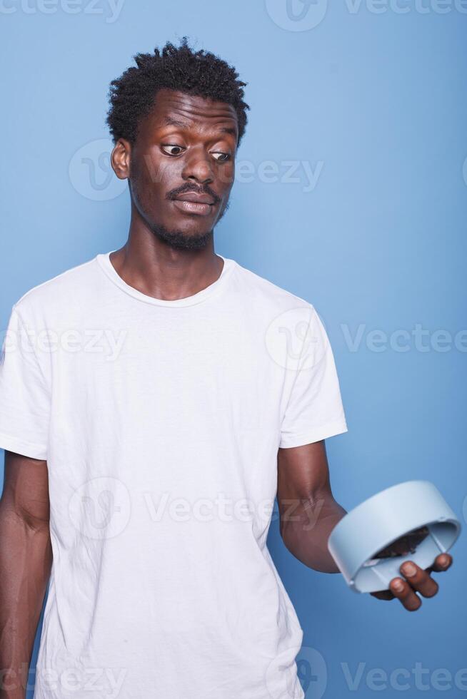 Close-up of african american man carrying wall clock with shocked expression. Portrait of black male individual with surprised face, looking at hours and minutes on a blue watch for alarm. photo