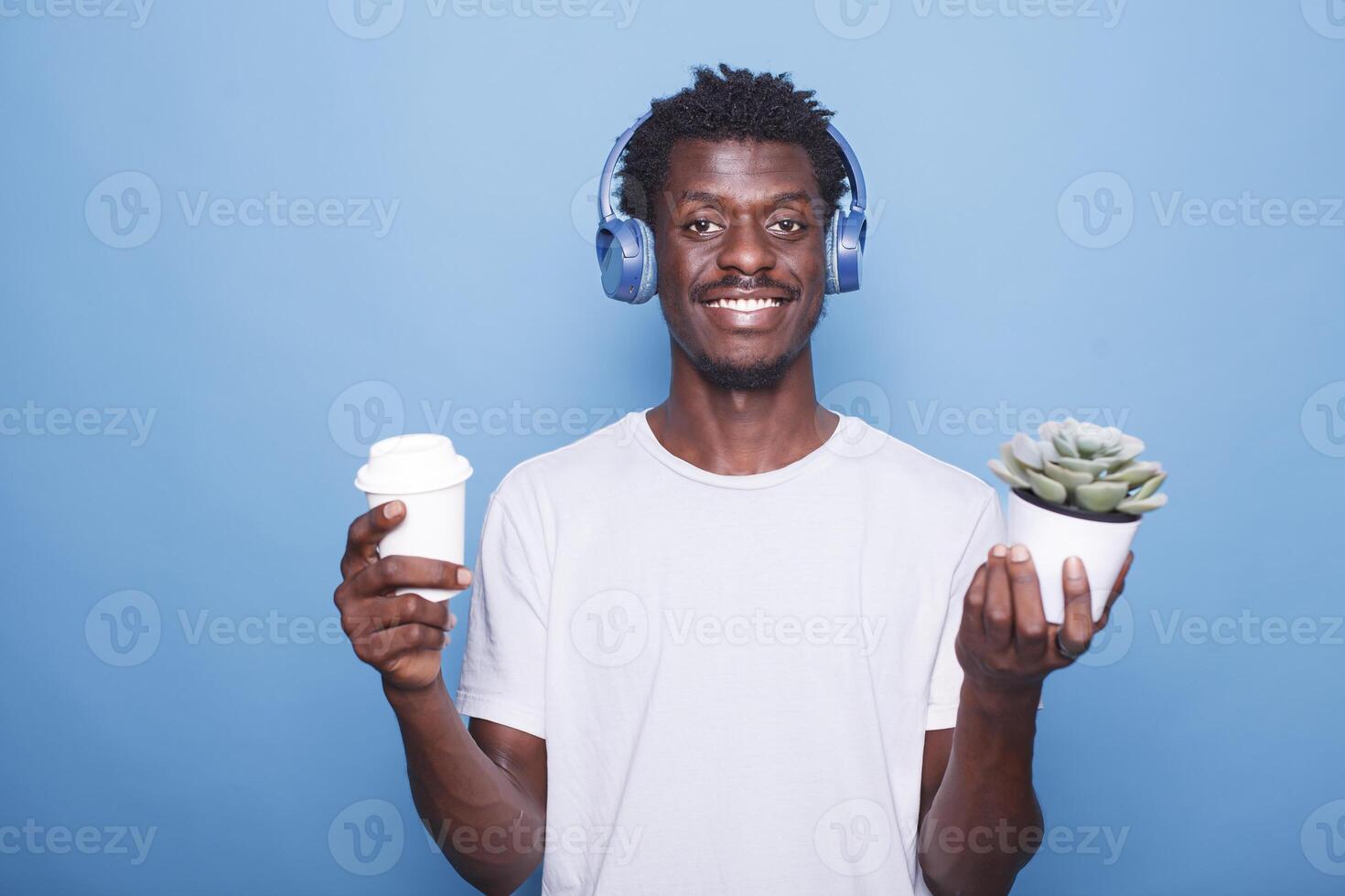 Portrait of happy African American man standing in front of a blue background with a potted plant while carrying a beverage. Smiling black guy holding a coffee cup and wearing wireless headphones. photo