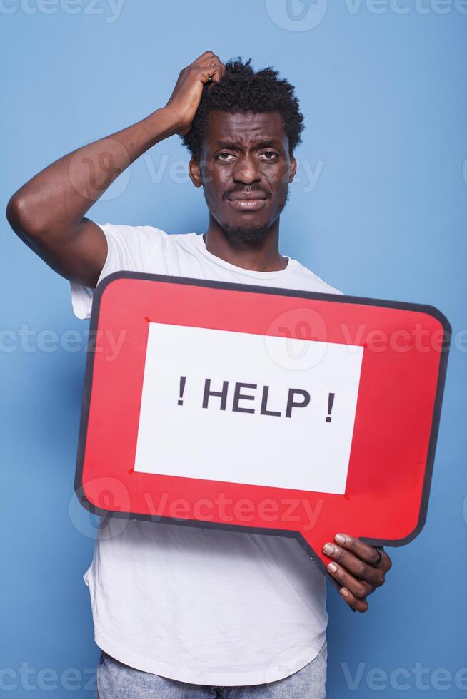 Bewildered african american individual holding voice bubble with text message asking for help. Portrait of black guy with hand on his head is grasping a red cardboard sign. photo