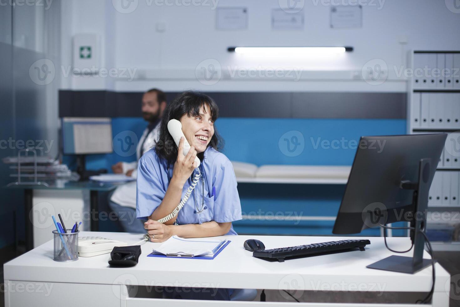 Smiling female practitioner sitting at the clinic office desk is talking on the landline. Caucasian nurse on phone call while using a desktop computer in a hospital room. photo