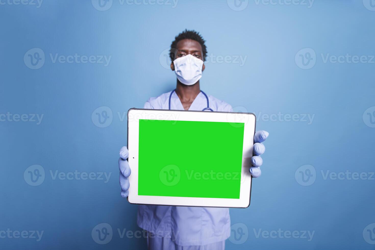 Male healthcare professional stands with a tablet displaying blank green screen while wearing scrubs and face mask. Holding digital device with chromakey template is a black man. photo