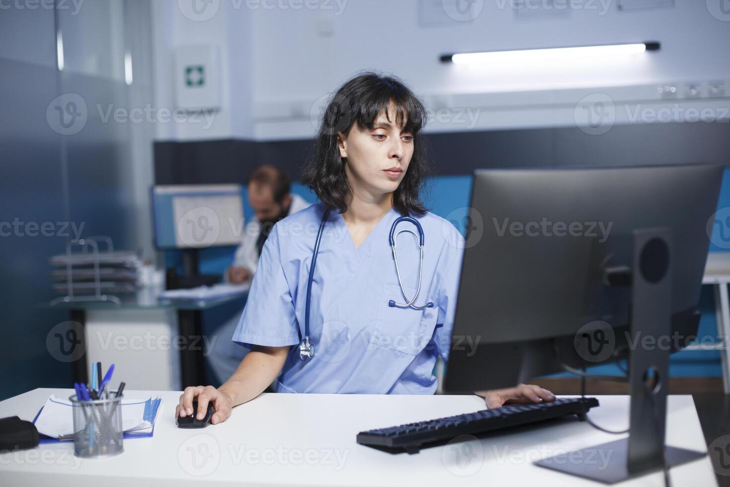 Detailed image of a caucasian nurse operating a desktop computer in a hospital office. A female practitioner wearing blue scrubs checks medical files on a computer in a clinic room. photo