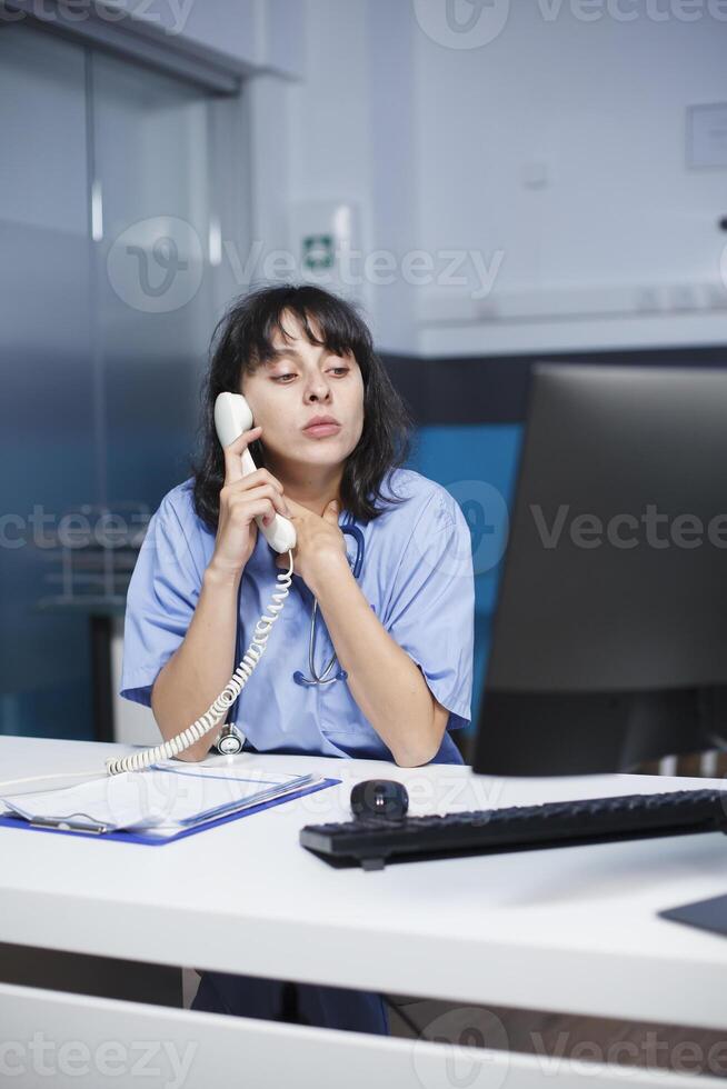 A medical nurse is on the telephone with a patient regarding his or her diagnosis. In a modern clinic, a female healthcare provider sits at a desk, using a landline phone and looking at a monitor. photo