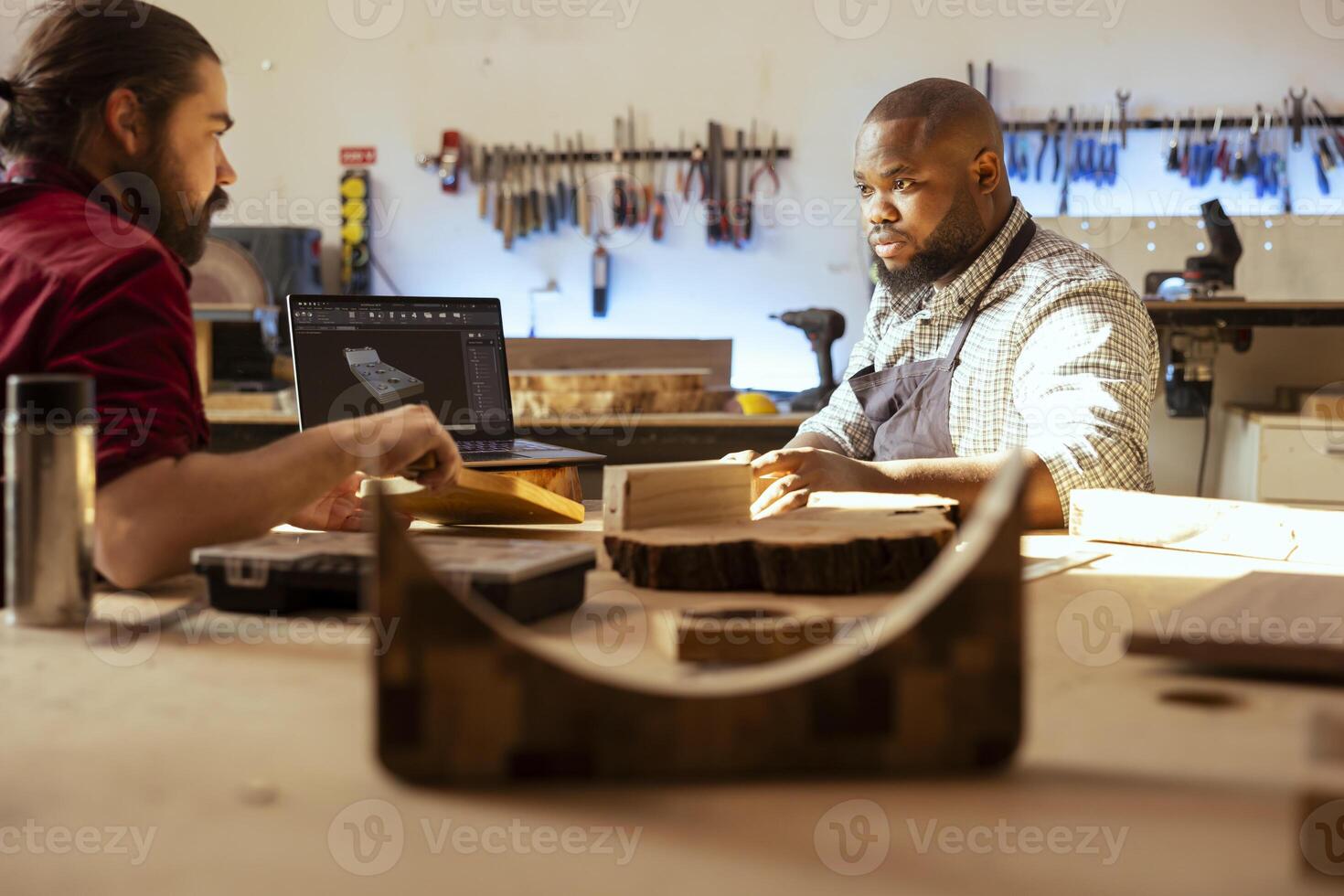 Cabinetmaker and colleague comparing wood piece used for furniture assembly with schematic displayed on laptop. Carpenter team in joinery crosschecking wooden object with notebook blueprint photo