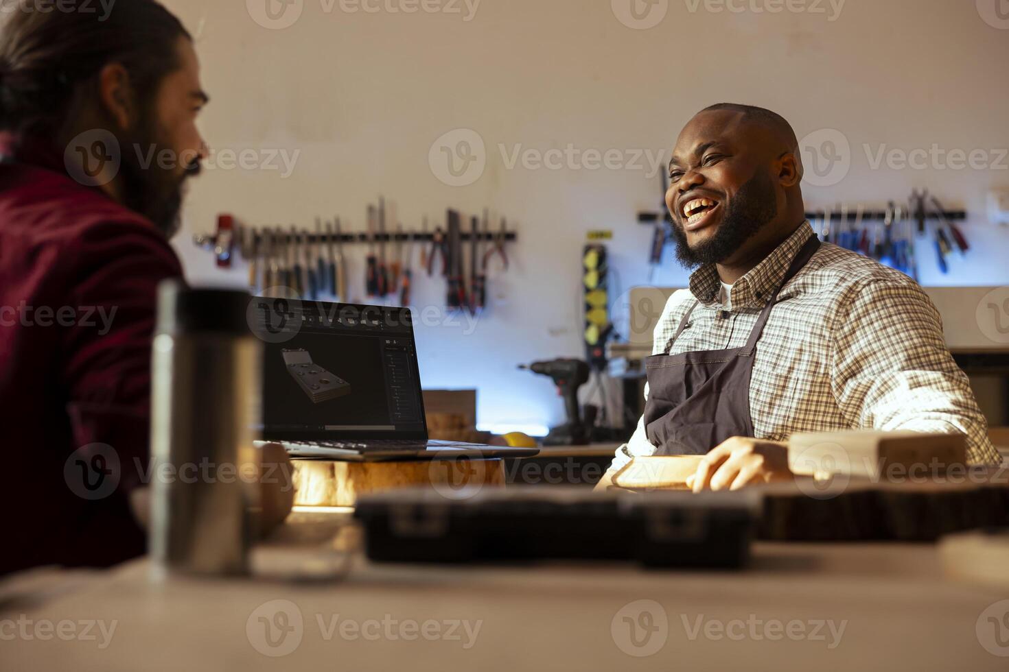 Happy carpenter looking over blueprints schematics on laptop in CAD software with cheerful apprentice. Smiling woodworker and joyful colleague having fun designing custom 3D furniture on notebook photo