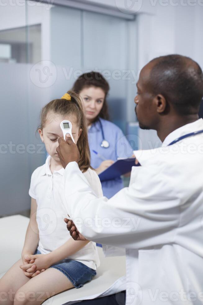 africano americano médico utilizando termómetro en pequeño niña a cheque fiebre en médico oficina. médico medidas el temperatura a encontrar un diagnóstico y da tratamiento a el niño en un gabinete. foto