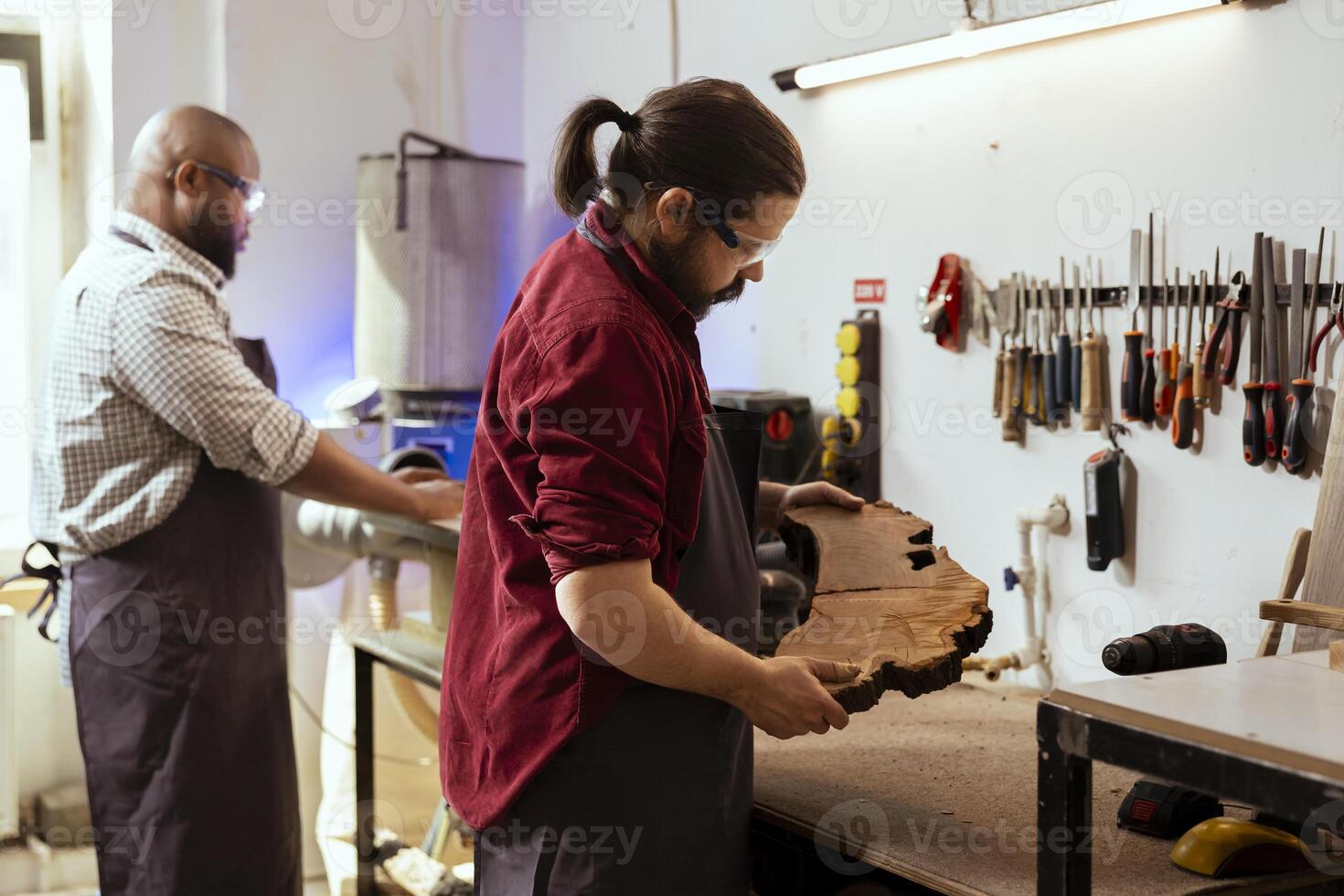 Carpenter holding timber block, doing quality assurance on it with coworker in background starting machinery. Manufacturer in joinery preparing piece of wood next to african american apprentice photo