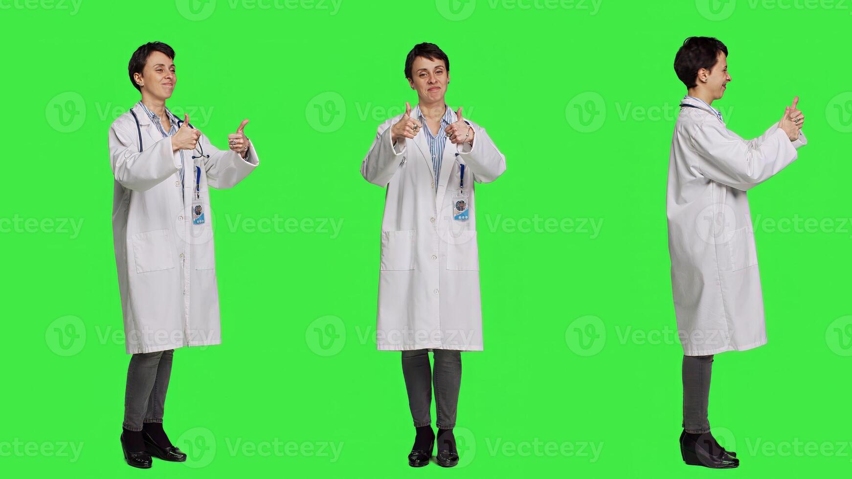 Cheerful medic doing thumbs up symbol against greenscreen backdrop, expresses positivity with like sign. General practitioner with coat giving approval and being satisfied with success. Camera B. photo