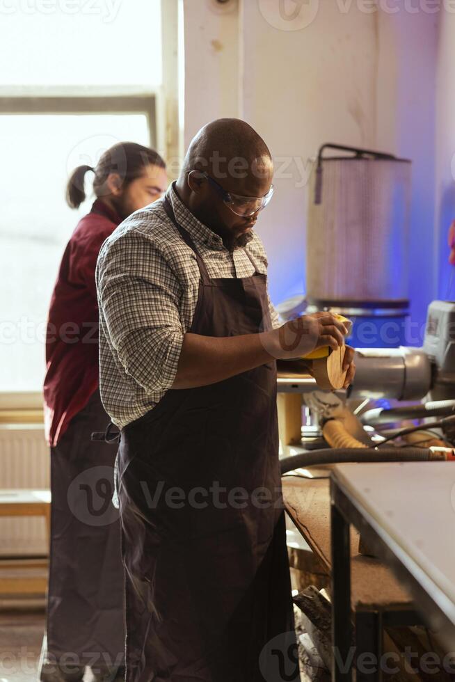 African american artisan in studio using sandpaper for smoothing wooden surface, creating wood art designs. BIPOC person using sanding sheets to refurbish damages suffered by timber block photo
