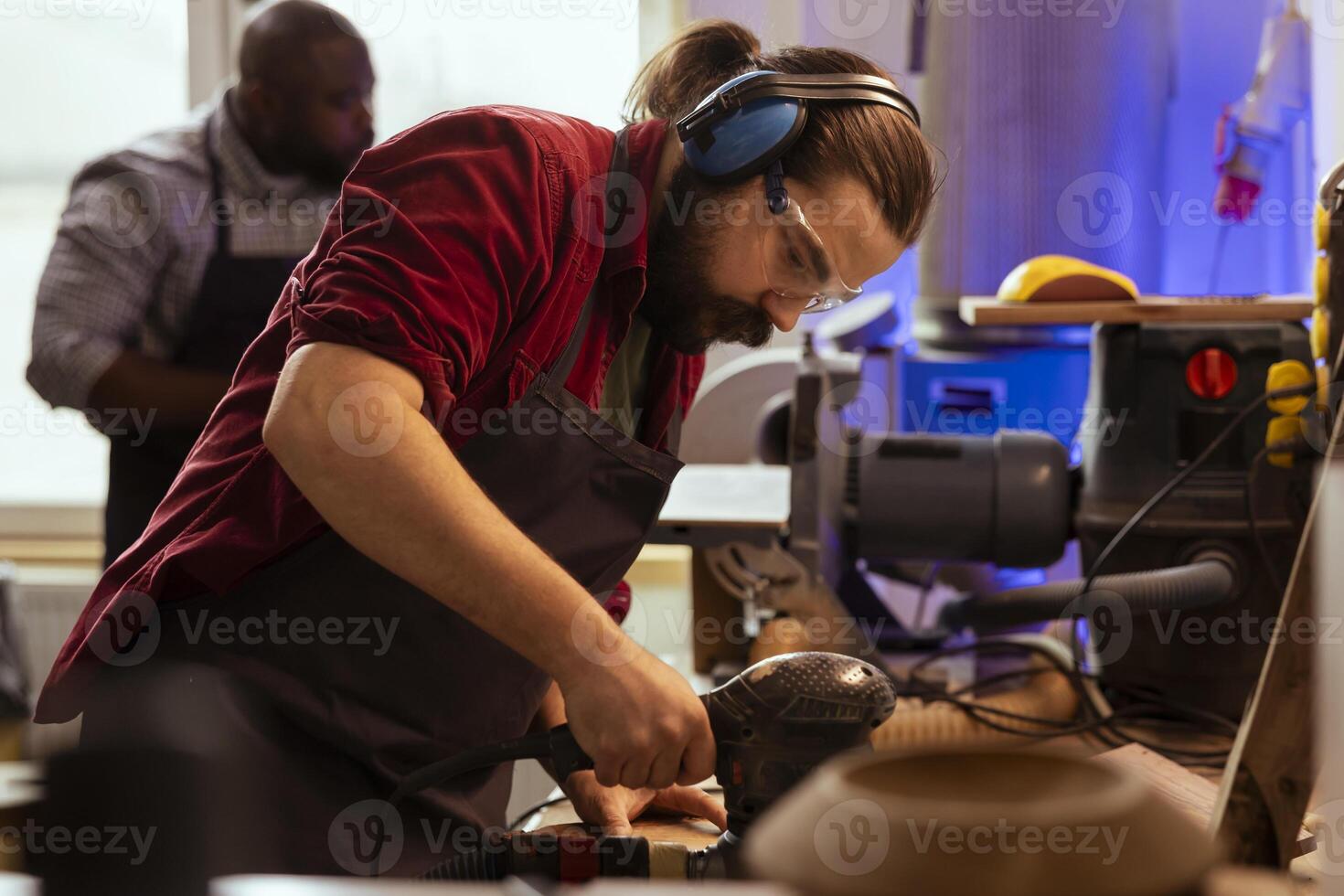 Artisan in joinery wearing safety earmuffs using orbital sander on lumber. Man in carpentry shop uses angle grinder to create wooden objects helped by colleague in blurry background photo