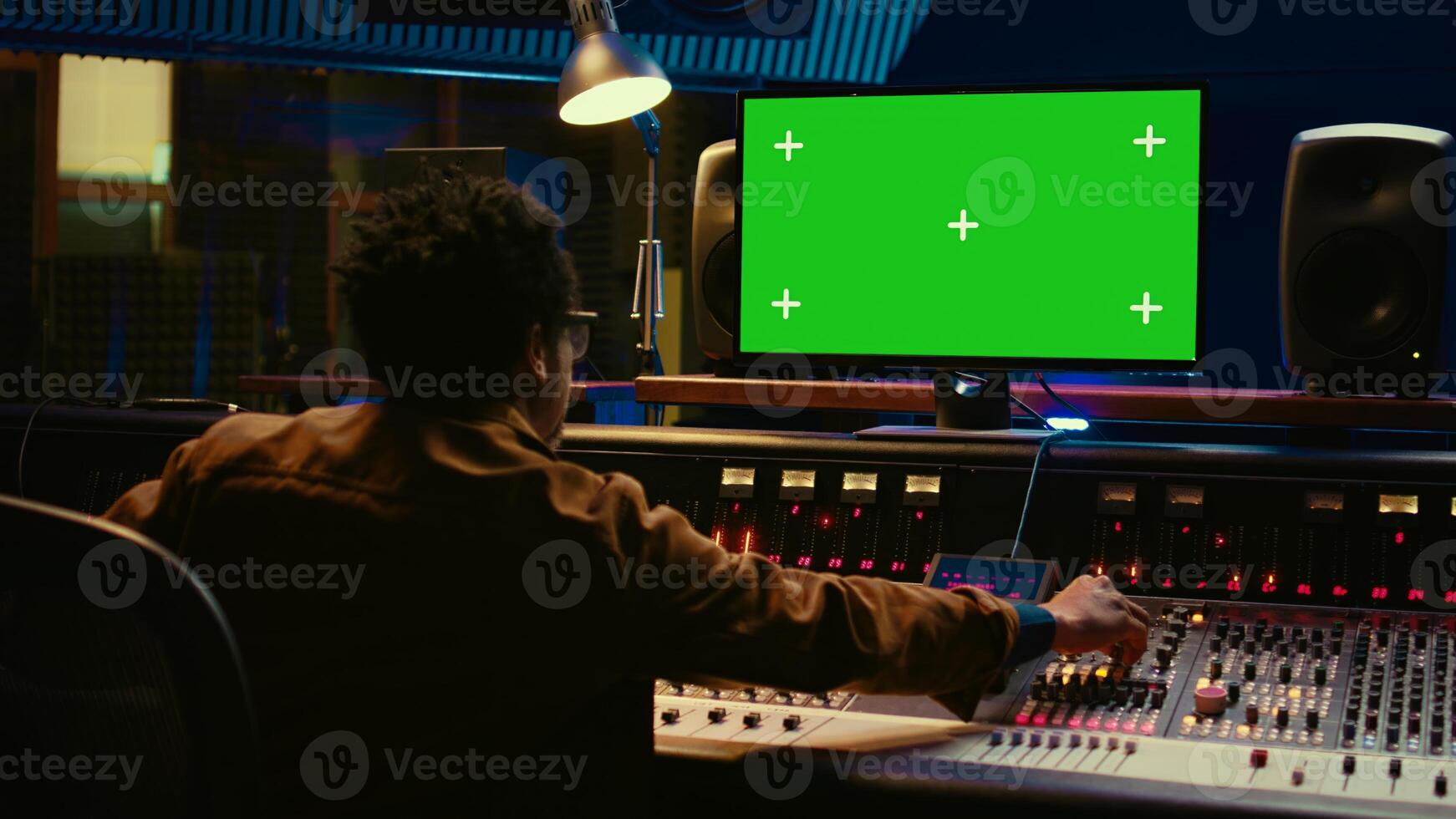 African american audio engineer uses greenscreen on pc in professional recording studio, working with mixing console and twisting knobs to edit tracks. Music producer creating tunes. Camera B. photo