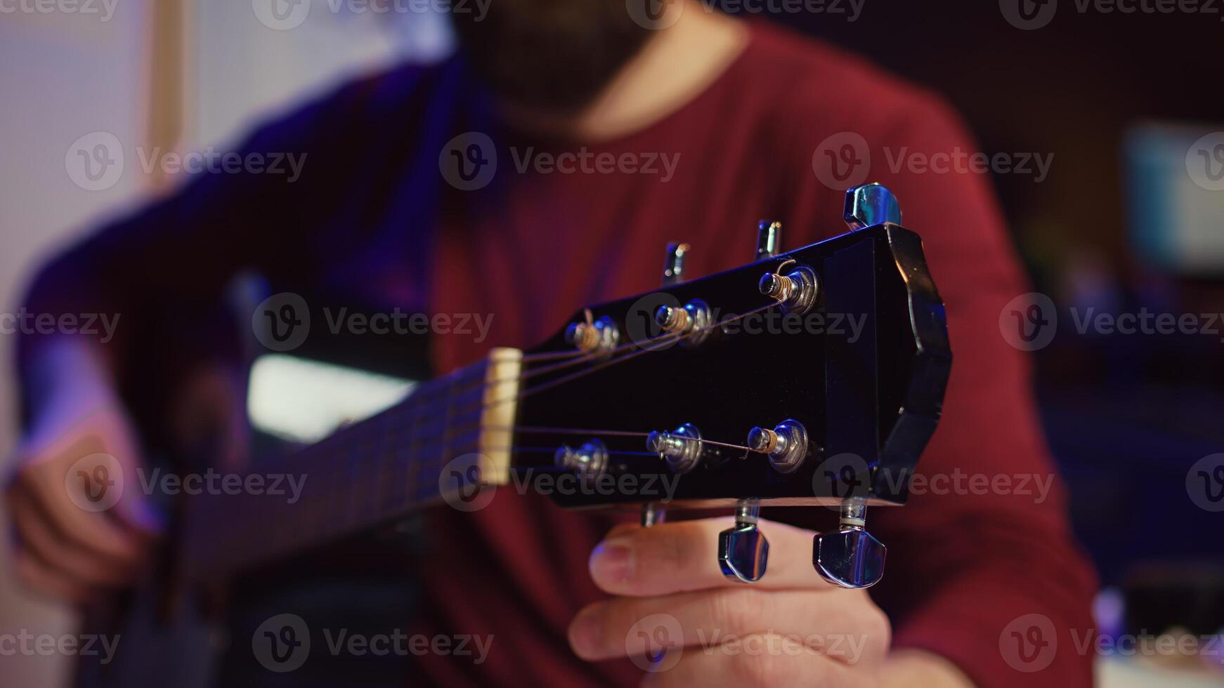 Musical performer tuning his guitar by twisting the knobs, preparing to play acoustic instrument in home studio. Artist recording sounds to create new track, using equalizer mixing gear. Camera B. photo