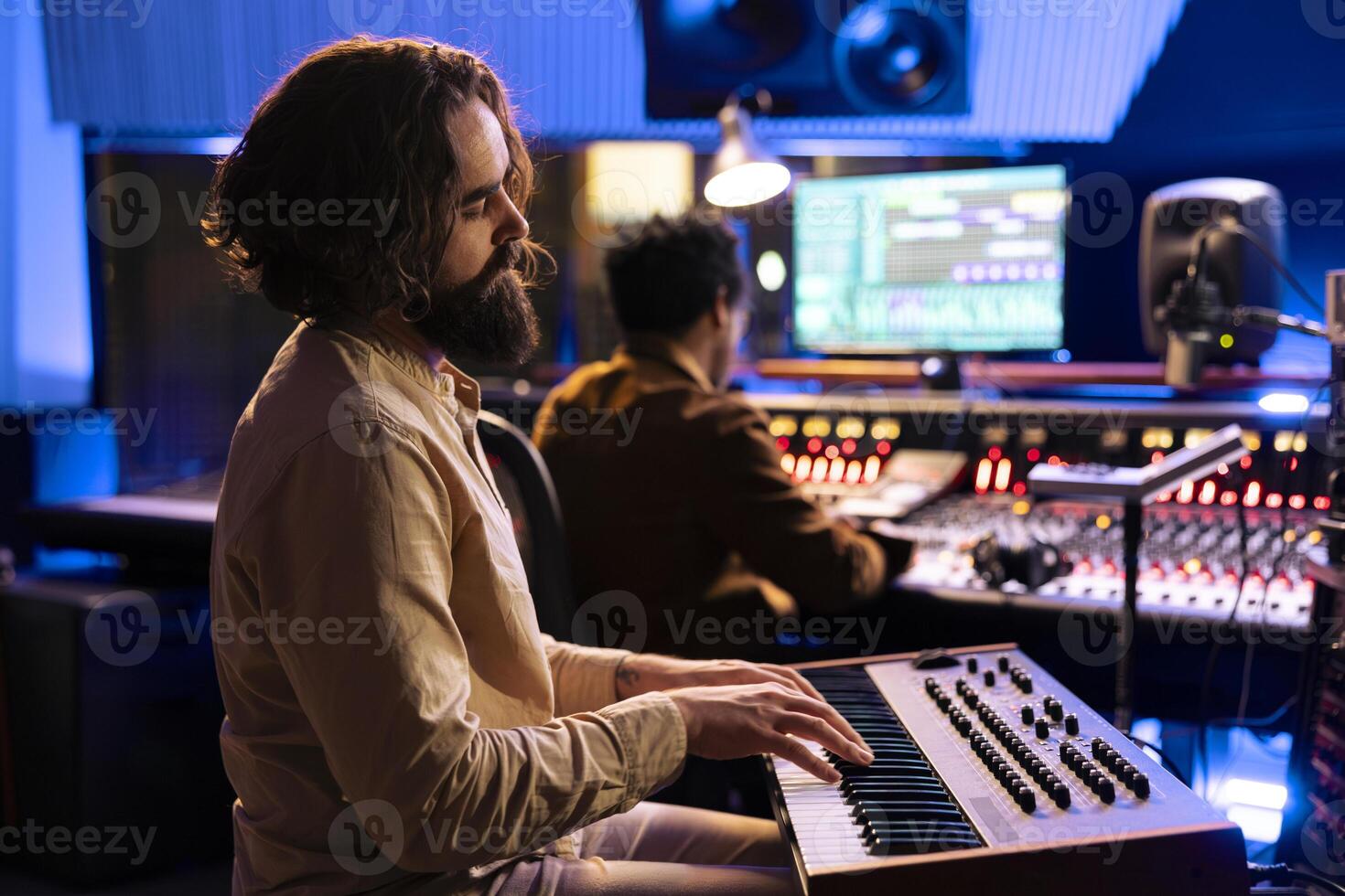 Young artist songwriter creating a new song with piano keys in studio, collaborating with audio engineer in control room. Musician singer composing music with midi controller electronic keyboard. photo