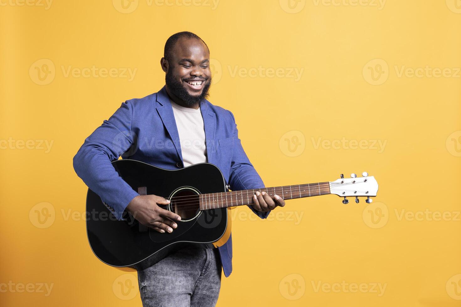 Smiling singer holding guitar, performing blues tunes during concert isolated over yellow studio background. Merry musician performing composition on stringed musical instrument photo