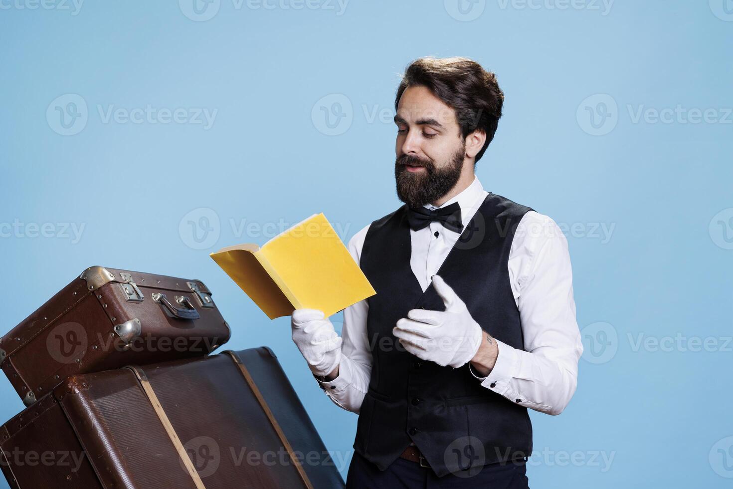 Stylish employee enjoys fiction book while on duty, taking care of trolley bags in studio. Male doorkeeper reading literature pages in front of camera, having fun with classic hobby. photo
