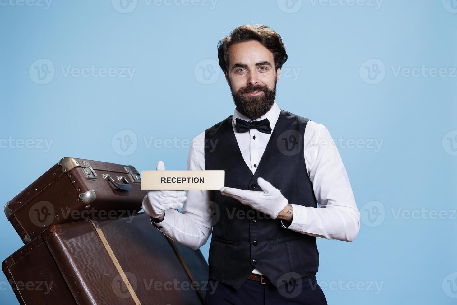 Hotel employee leading tourists in the right way using reception wall sign. Skilled professional bellboy posing on camera and holding front desk indicator, guiding guests and travellers. photo