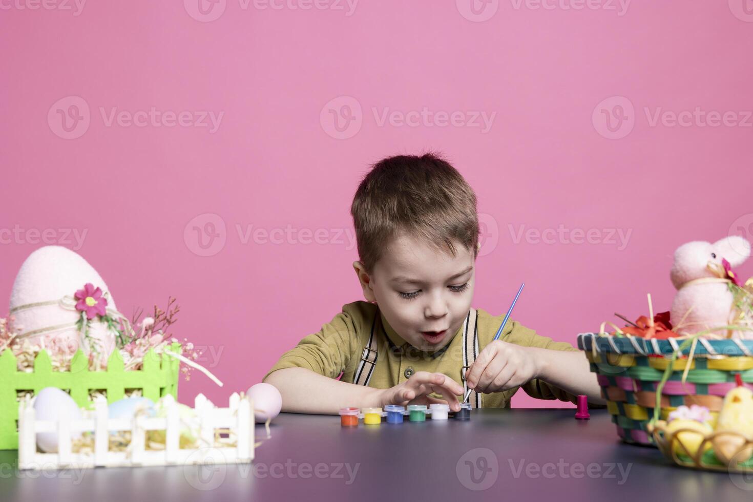 adorable niñito sonrisas como él usos acuarelas y pinturas a Decorar huevos y centros de mesa para el Pascua de Resurrección celebracion festividad. pequeño extático joven chico explorador creatividad con Arte suministros. foto