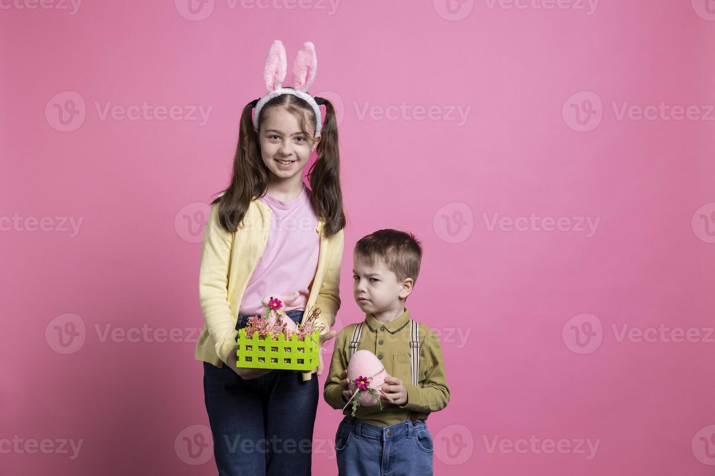 Playful children showing painted eggs and decorating for easter celebration festivity, standing together over pink background. Brother and sister feeling cheerful and enthusiastic about april event. photo